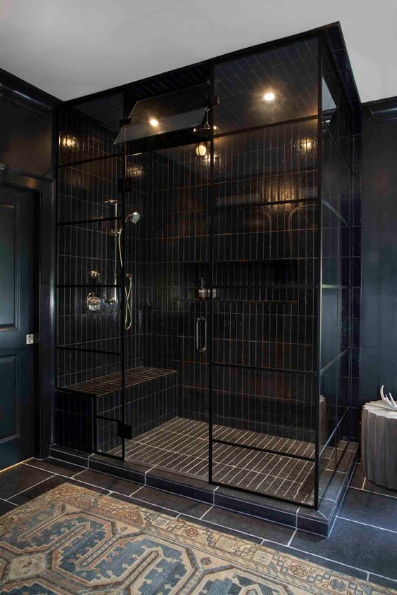a modern black bathroom clad with skinny and large scale tiles, a large shower space, a side table and a printed rug