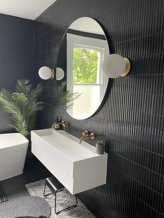 a modern black tropical bathroom with a skinny tile wall, white appliances, a round mirror with sconces and a potted tropical plant