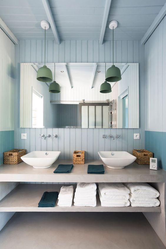 a modern light blue bathroom with beadboard, a concrete vanity and green pendant lamps over the sinks