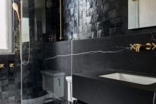 a modern moody bathroom with catchy tiles and black marble, a floating vanity, gold fixtures and a mirror