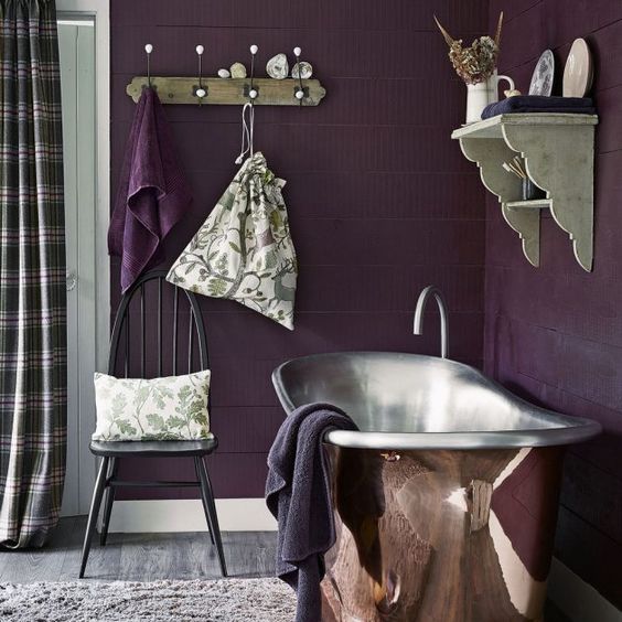 a moody rustic bathroom done in deep purple, with a metal tub, vintage shelves and a chair plus plaid curtains