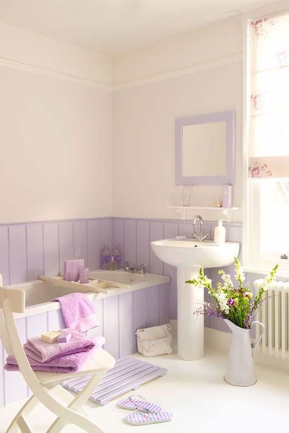 a romantic bathroom with lavender paneling and a mirror, a wooden mat and a vase plus purple towels