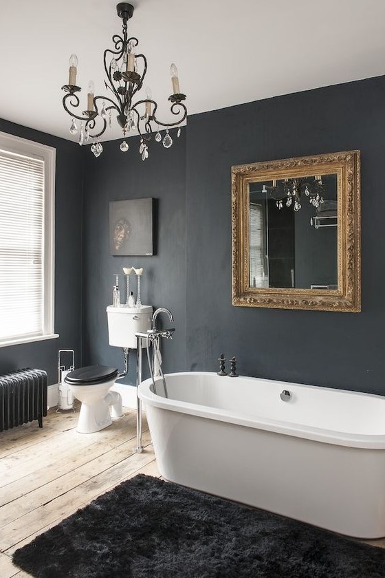 a vintage black bathroom with an oval modenr tub, a mirror in a chic gilded frame, a black rug, a black radiator and a crystal chandelier