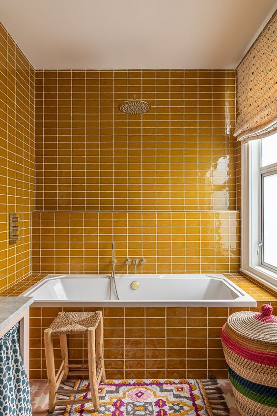a warm yellow bathroom fully clad with tiles, bright printed textiles, a basket with a lid for storage and a window with frosted glass