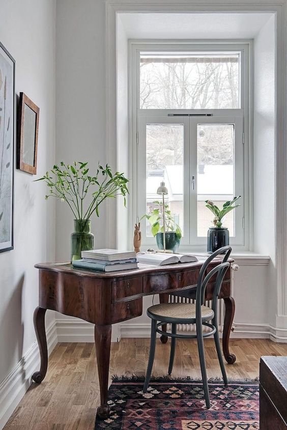 a beautiful vintage home office space with a large heavy stained desk, a grey chair, some potted plants and large books