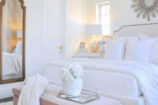a chic glam bedroom in white with an upholstered bed and bench, a vintage mirror, a crystal chandelier and a sunburst mirror