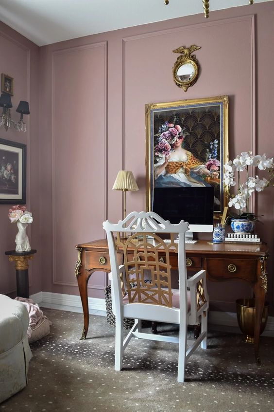 a gorgeous vintage working space with mauve walls, a stained vintage desk, a white and gold chair, a bold artwork and some gold decor