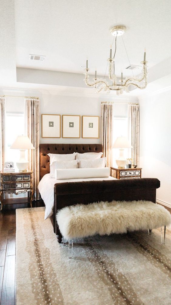 a modern glam bedroom with a brown bed, a crystal chandelier, mirror nightstands, a faux fur bench and printed textiles
