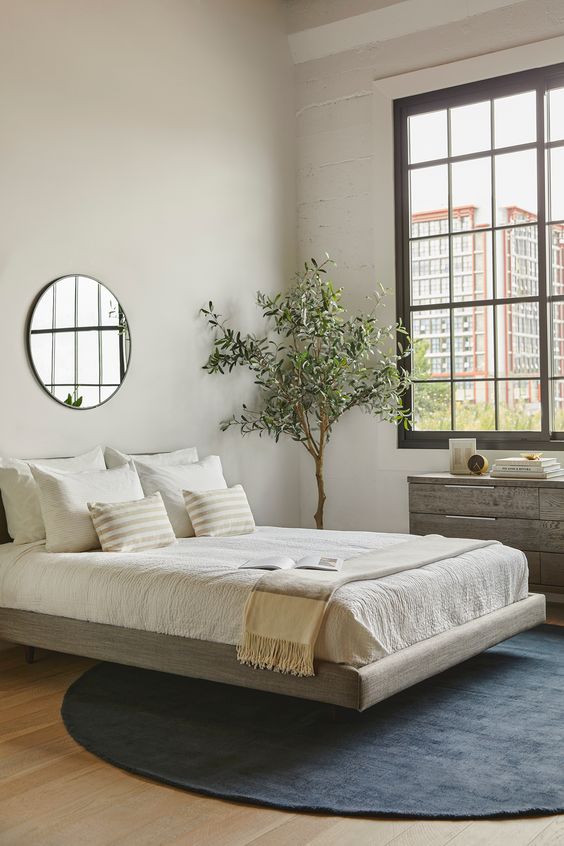 a modern zen bedroom with a grey floating bed, a reclaimed wooden dresser, a statement plant, neutral bedding and a round mirror