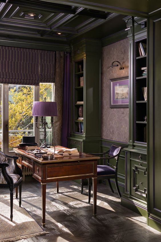 a moody vintage home office with grene cabinetry, mauve floral wallpaper, a stained desk and vintage chairs, purple accessories