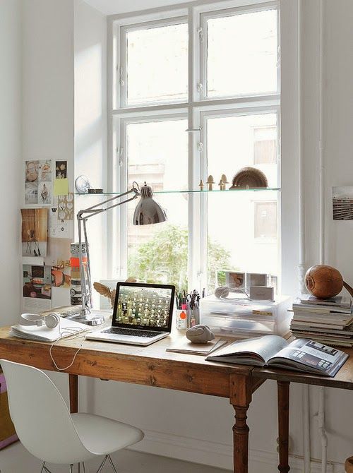 a pretty vintage home office nook by the window, with a stained desk, a white chair, a table lamp, a window shelf and some posters
