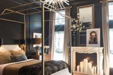 a refined dark glam bedroom with black walls, a faux fireplace, a gold bed, a large mirror and a statement chandelier