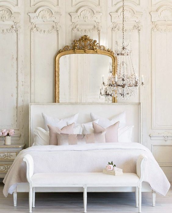 a refined glam bedroom with molding on the walls, a statement mirror, a crystal chandelier and upholstered furniture