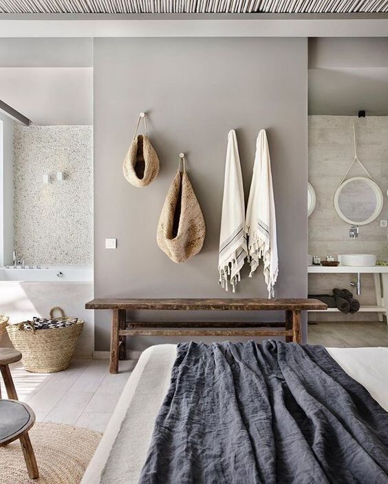 a relaxed zen bedroom united with a bathroom, with stone and concrete walls, a wooden bench, neutral bedding and baskets for storage