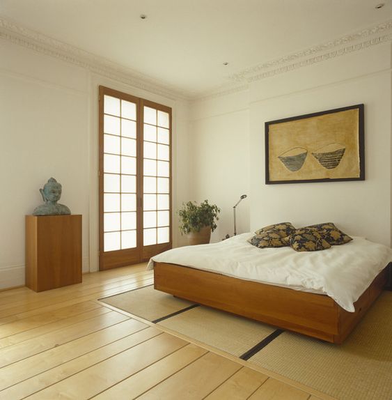 a relaxed zen bedroom with a wooden bed and a stand with a statue, a statement plant, neutral bedding and a bold artwork