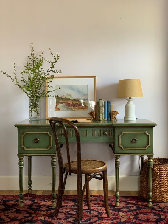 a small and chic vintage working space with a green desk, a stained chair, a table lamp, some books and greenery in a vase