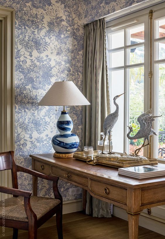 a sophisticated vintage working space with blue floral wallpaper, a stained desk, a stained chair, a gold tray with decor and a table lamp