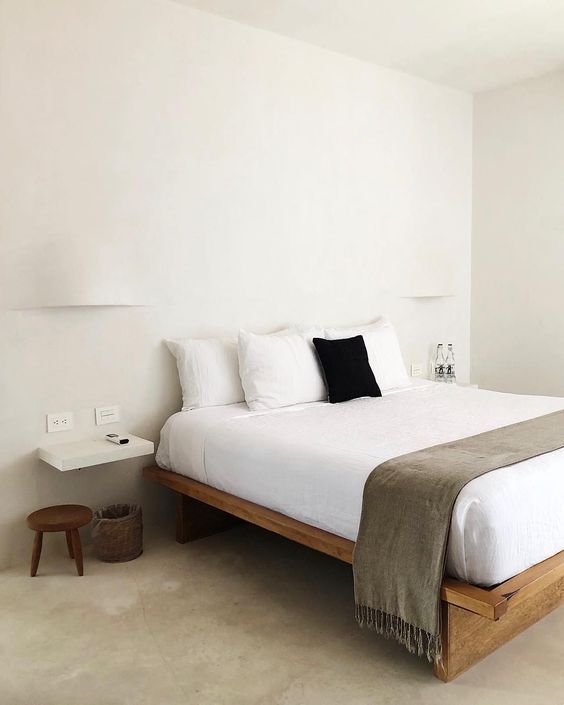 a very simple zen bedroom with a simple wooden bed, a low stool and floating nightstands, neutral bedding and wall lamps