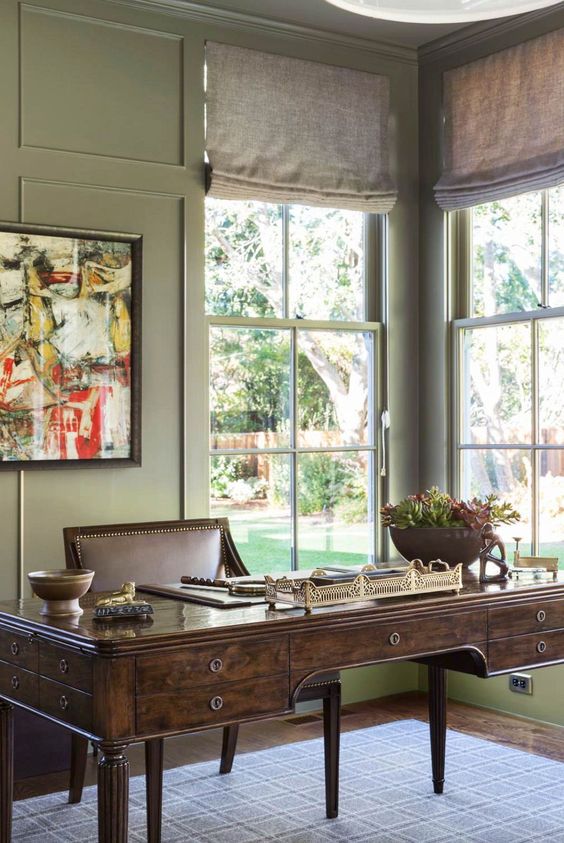 a vintage home office with green walls, a dark-stained vintage desk and a brown leather chair, neutral curtains and a bold artwork