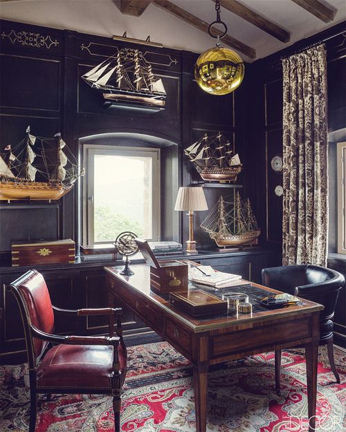 a vintage moody home office with black walls and cabinetry, a stained desk and a black and a red chair, ships for decor and a gold bubble lamp