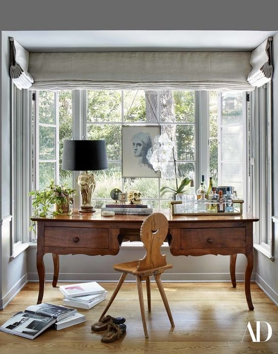 a vintage working nook by the window, with a large stained desk and a light-stained chair, a vintage table lamp and some art