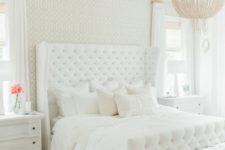 a white glam bedroom with a statement upholstered bed, white and floral furniture, a fringe chandelier