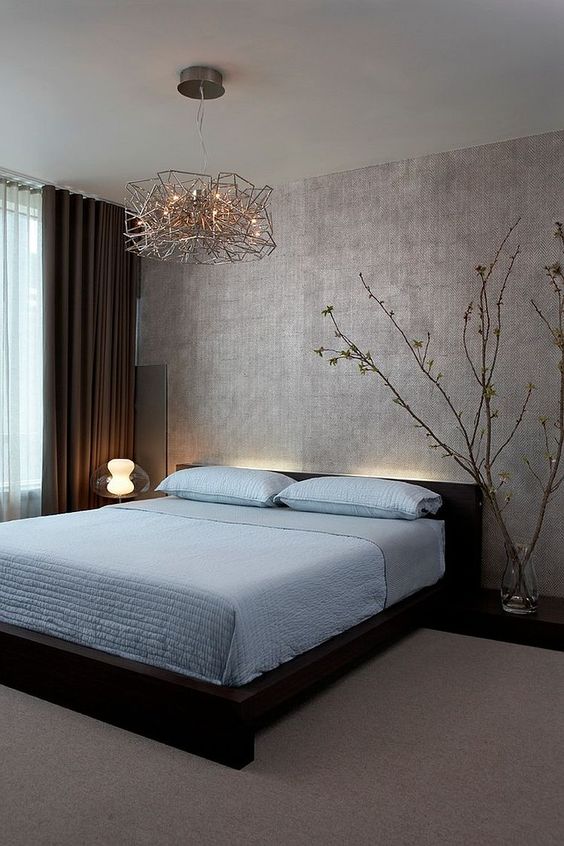 a zen-like bedroom with a silver accent wall, a dark bed with blue bedding, blooming branches and very eye-catchy lamps