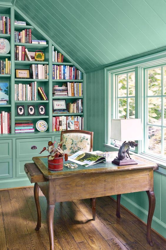 an attic home office with green walls and a ceiling, a built-in bookcase and a stained desk, a floral chair and a table lamp