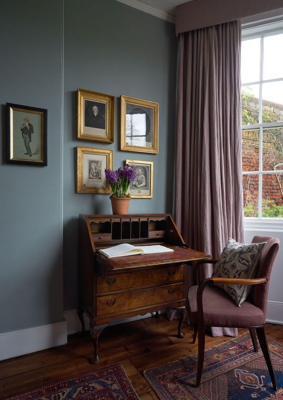an exquisite vintage working nook with grey walls, a vintage dark-stained bureau and a mauve chair, mauve curtains and a gallery wall