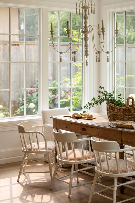 a French cottage dining room with a stained table, vintage white chairs, a chic chandelier and a basket with greenery