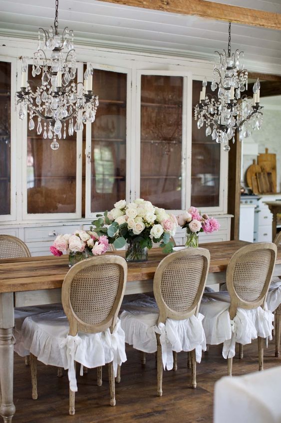 a French country dining room with several white buffets, a stained rustic table, vintage cane chairs and crystal chandeliers