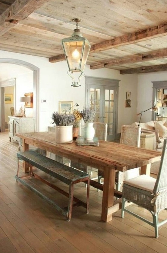 a Provence dining room with a wooden floor and ceiling with beams, a stained table, benches and woven chairs, a cool pendant lantern