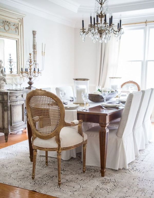 a beautiful neutral French dining room with a stained table and neutral chairs, a vintage storage cabinet, a mirror and a crystal chandelier