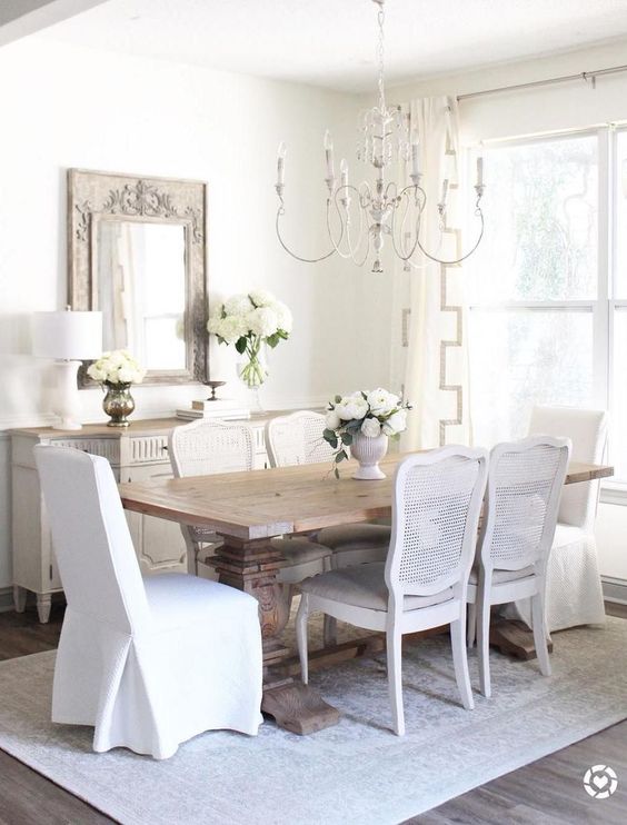 a chamring French country dining room with a neutral sideboard, a stained vintage table, white chairs, a lovely chandelier and a mirror in a wooden frame