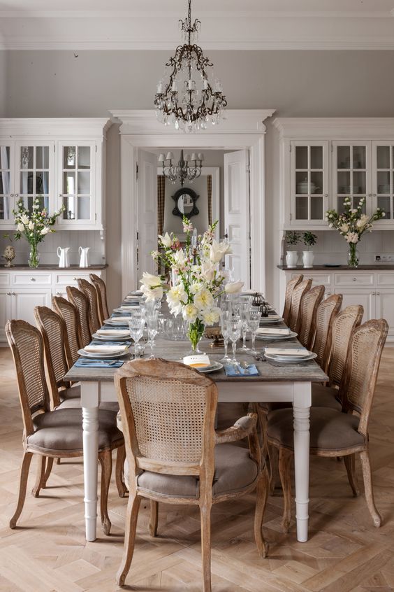 a chic French dining room with grey walls, white cabinetry, a large table and cane chairs, a crystal chandelier