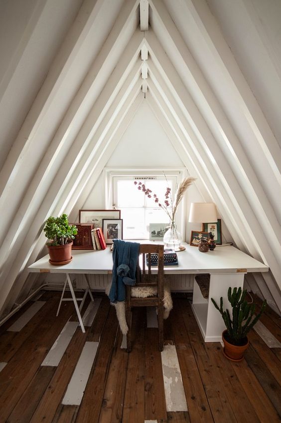 a cool attic home office with a desk by the window, a vintage chair and a reclaimed wood floor, potted greenery and artworks