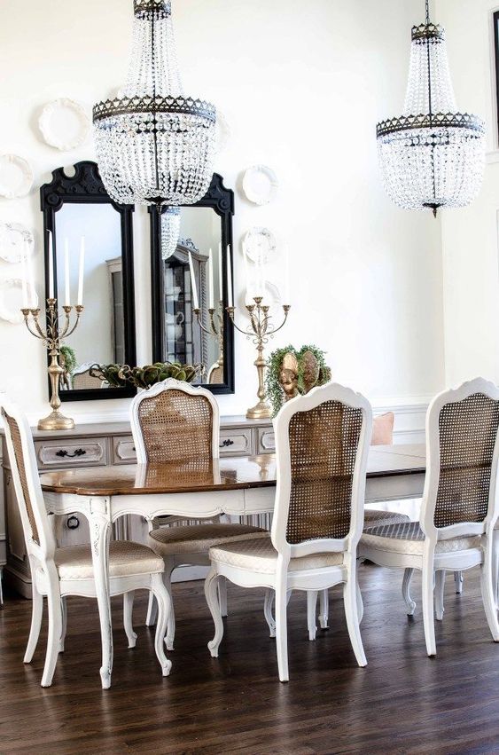 a gorgeous vintage French dining room with a whitewashed sideboard, an oval table and vintage cane chairs, lovely chandeliers and mirrors in black frames