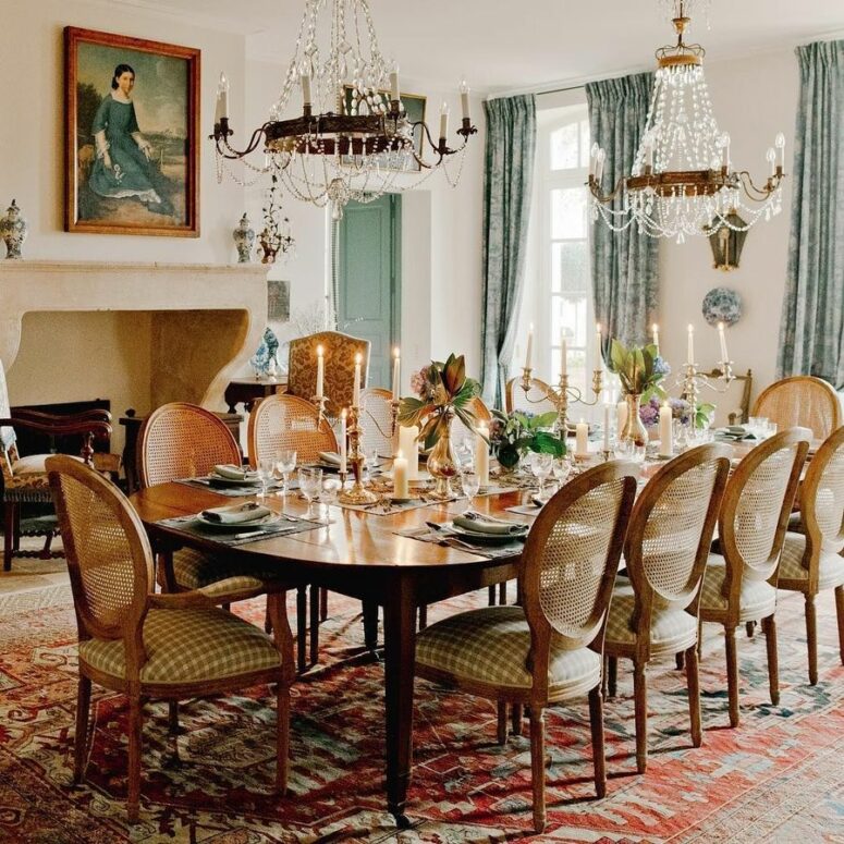 a jaw-dropping vintage French dining room with a non-working fireplace, a large stained table and vintage chairs, crystal chandeliers and a vintage artwork