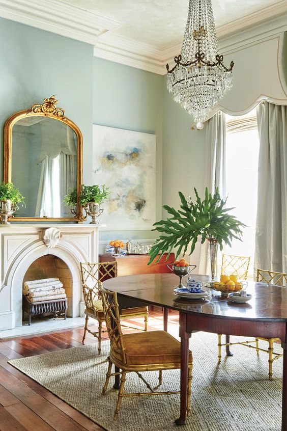 a lovely French chic dining room with light blue walls, a non-working fireplace, a stained table and vintage chairs, a large crystal chandelier