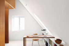 a minimalist attic home office with a long bookshelf and a built-in desk, a chic table lamp and a comfy leather chair is filled with light and chic