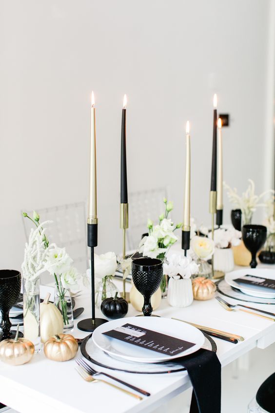 a minimalist monochromatic Halloween tablescape wth black, gold and neutral pumpkins, candles, black napkins and goblets, white blooms