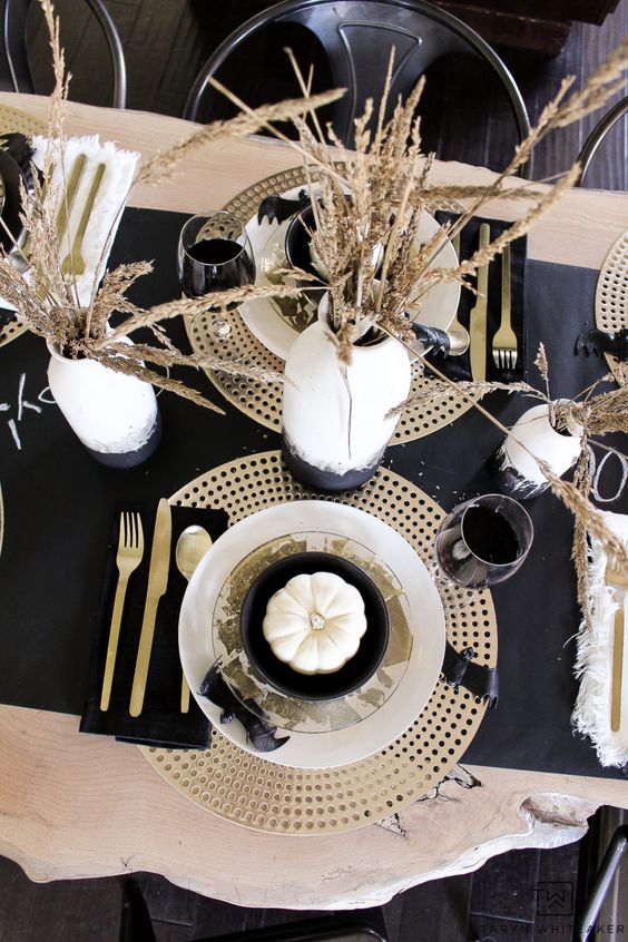 a modern glam Halloween table setting with gold chargers, cutlery, a black runner and watercolor vases with wheat