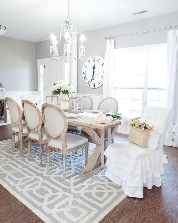 a neutral French country dining room with grey walls, white paneling, a rustic dining table, vintage neutral chairs, a ruffle chair cover on one of them and a crystal chandelier