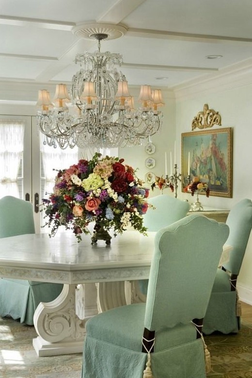 a neutral French dining room with a large stone tabletop table, mint green chairs, a large crystal chandelier, a large artwork