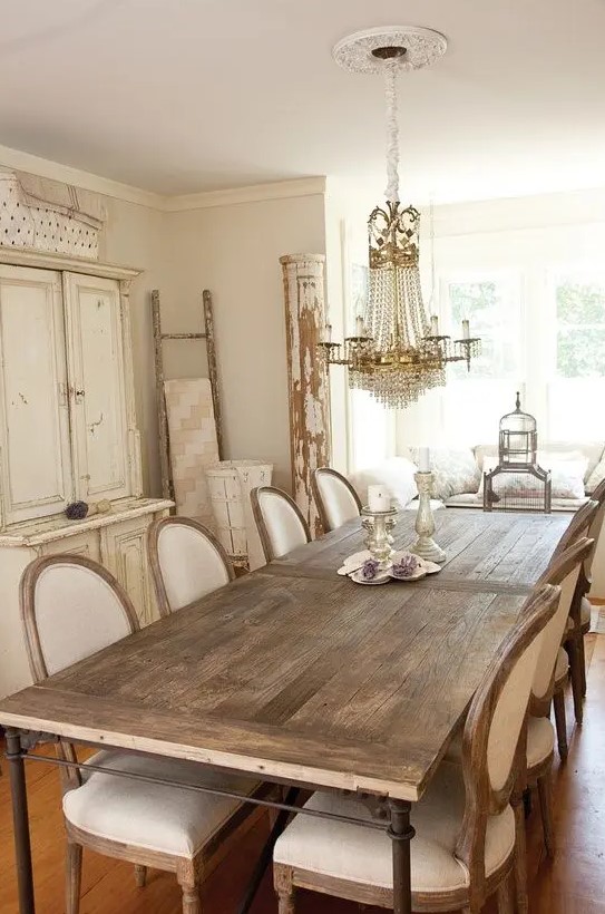 a neutral French dining space with a large storage unit, a stained table, white vintage chairs, a crystal chandelier and some vintage decor