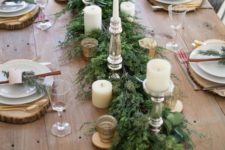 a simple and elegant winter table with a greenery runner, candles, wood slice placemats, cinnamon bark, evergreens and gold touches