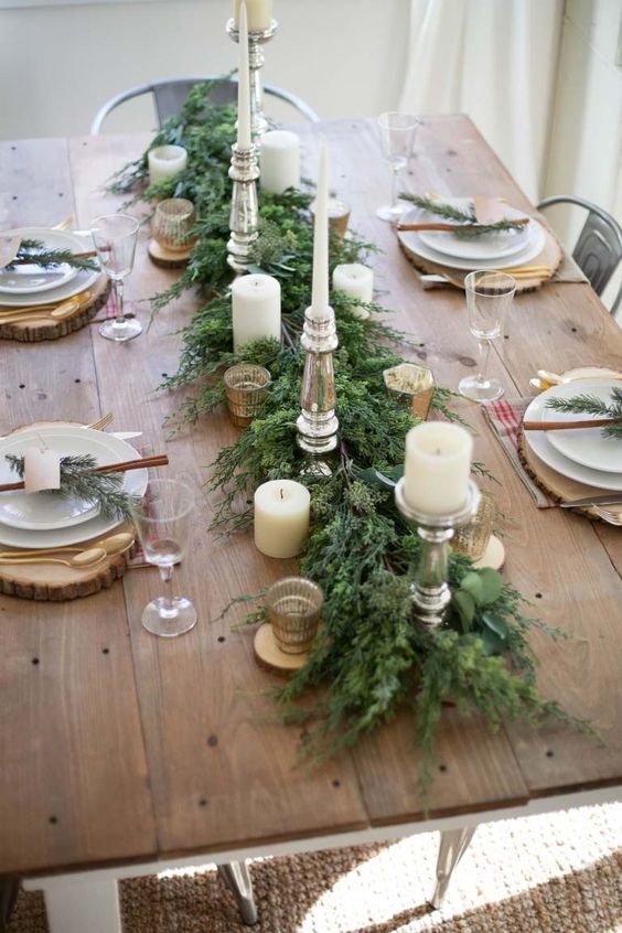 a simple and elegant winter table with a greenery runner, candles, wood slice placemats, cinnamon bark, evergreens and gold touches