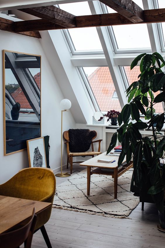 a small and elegant attic home office with a reading nook by the window, a wooden chair and a low coffee table, a desk and a mustard chair plus lots of skylights