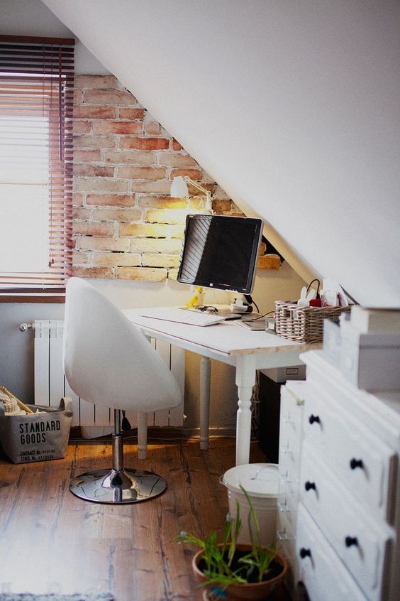 a small attic home office with a vintage desk and dresser, a white egg-shaped chair, potted greenery and a brick accent wall