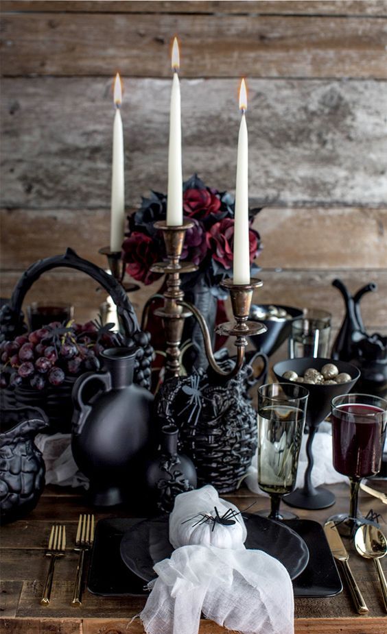 a sophisitcated Halloween tablescape with deep red blooms, fruit, black vases and jugs, candles, white pumpkins and cheesecloth
